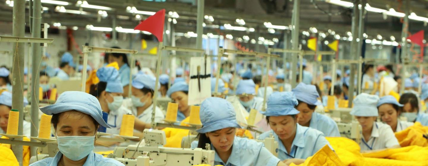 Operárias de uma fábrica têxtil usam uniformes azuis e máscaras enquanto operam máquinas de costura para produzir peças de roupa amarelas.