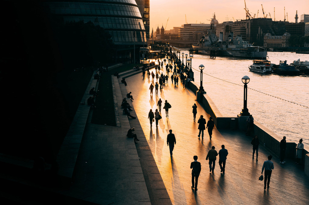 Silhuetas de pessoas caminhando ao longo de um calçadão à beira do rio em Londres ao pôr do sol.
