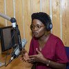 Mwanamke katika studio ya redio akizungumza kwenye kipaza sauti huku akiwa amevaa vipokea sauti vya masikioni.