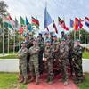 چھلاورن کی وردیوں اور نیلے رنگ کے ہیلمٹ میں UNIFIL امن فوجیوں کا ایک گروپ متعدد بین الاقوامی جھنڈوں والی یادگار کے سامنے تصویر کے لیے پوز دے رہا ہے۔