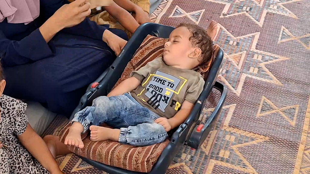 طفل صغير ينام في مقعد السيارة على سجادة منقوشة، محاطاً بأشخاص آخرين.