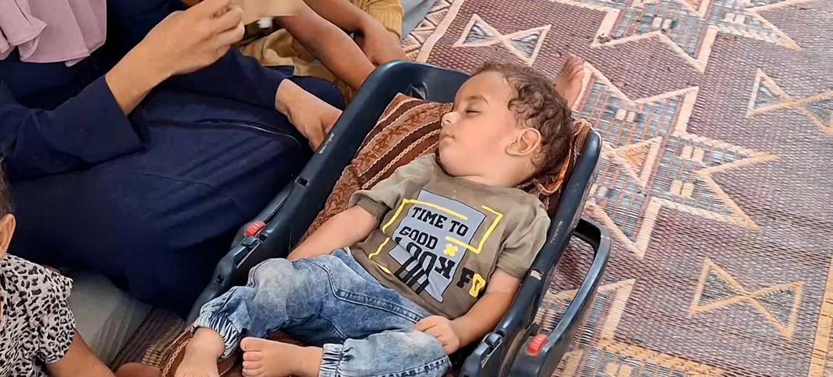 طفل صغير ينام في مقعد السيارة على سجادة منقوشة، محاطاً بأشخاص آخرين.
