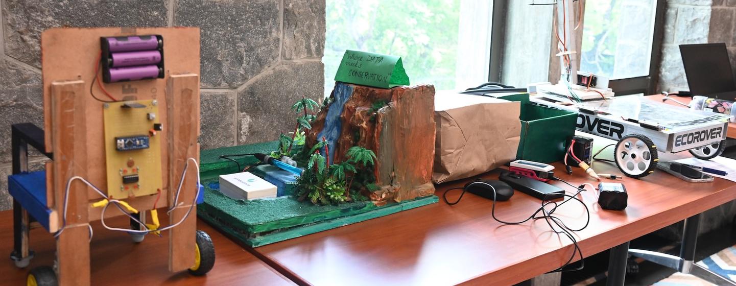 UNIC इंडिया के एक कार्यक्रम में छात्रों द्वारा निर्मित परियोजनाएं, जिनमें इलेक्ट्रॉनिक्स से सुसज्जित एक लकड़ी की गाड़ी और एक लघु भूदृश्य मॉडल शामिल हैं, एक मेज पर प्रदर्शित की गई हैं।