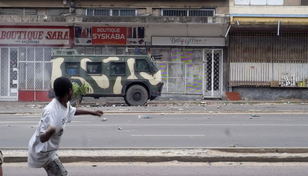Mvulana akitupa kitu barabarani huko Maputo, Msumbiji, huku gari la polisi lililofunikwa kwa silaha likiwa limeegeshwa mbele ya maduka nyuma.