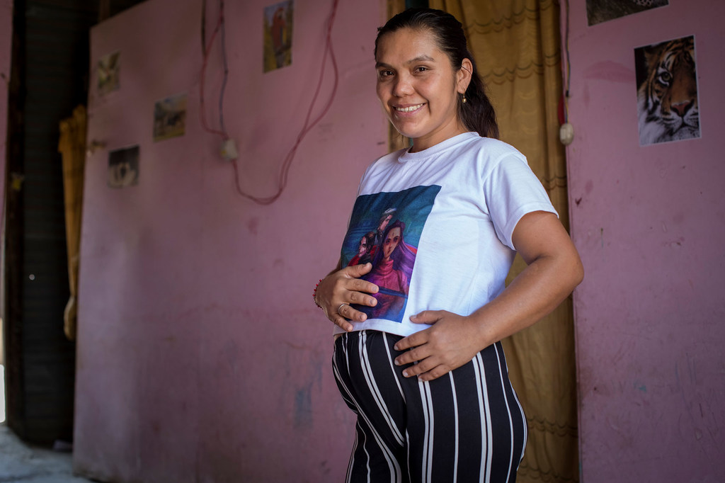 Una mujer embarazada, vestida con una camiseta blanca y pantalones a rayas, está de pie en el interior de la habitación, sonriendo mientras se toca la barriga. Las paredes son rosas y hay un póster de un tigre.