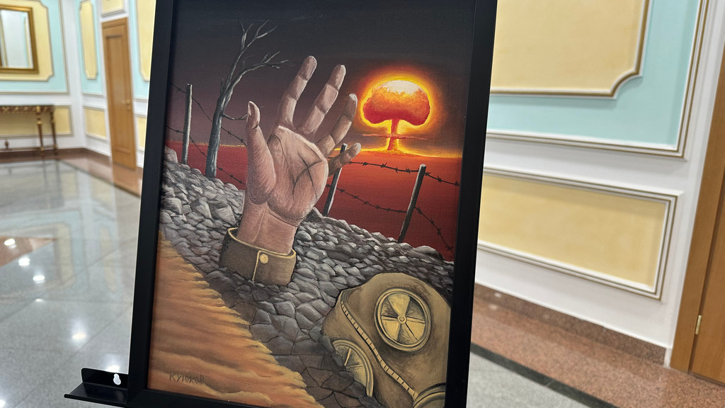 Une peinture d'Izumi Nakamitsu représentant une main sortant d'une tranchée remplie de fil de fer barbelé, avec un masque à gaz à proximité et un nuage en forme de champignon en arrière-plan, affichée dans une galerie.