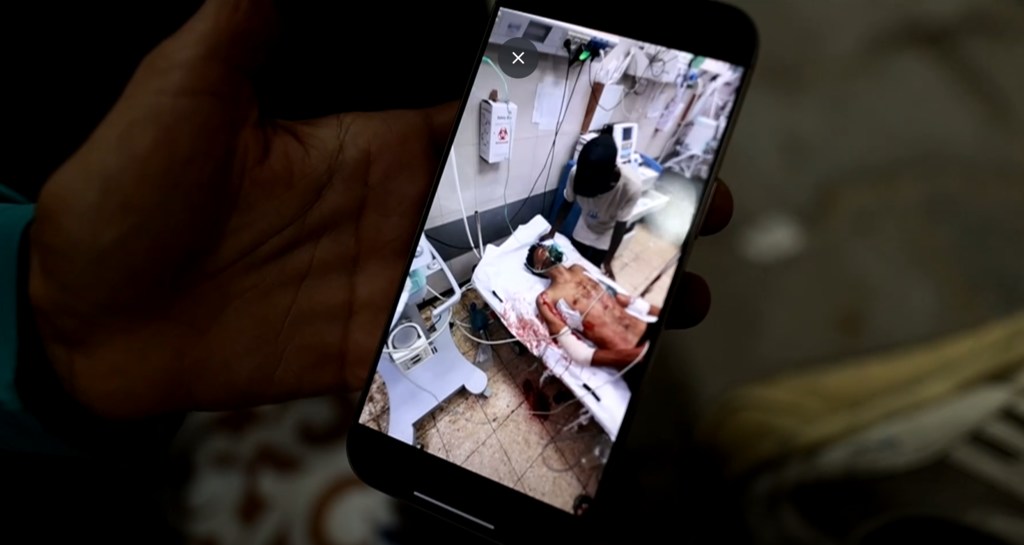 Una persona sostiene un teléfono inteligente que muestra la foto de un paciente ensangrentado tendido en una cama de hospital.
