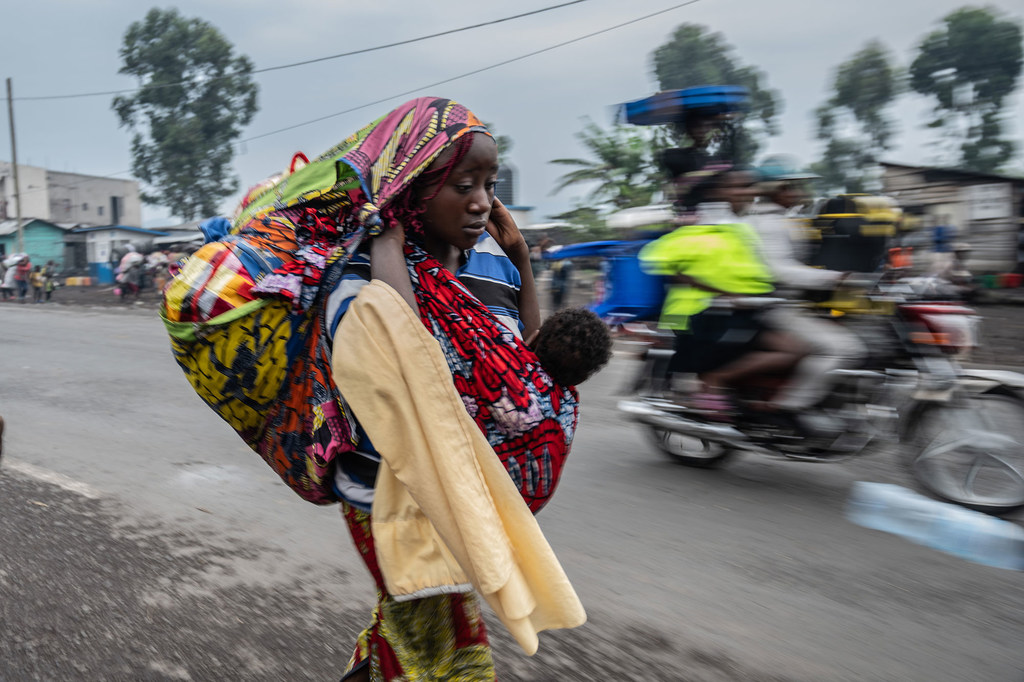 Mwanamke anayebeba mtoto mgongoni mwake na kifurushi kikubwa anatembea barabarani karibu na Goma, Jamhuri ya Kidemokrasia ya Kongo, wakati pikipiki inapopita.