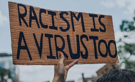 Une main brandit une pancarte en carton où l'on peut lire, au marqueur noir, le message « LE RACISME EST AUSSI UN VIRUS ».