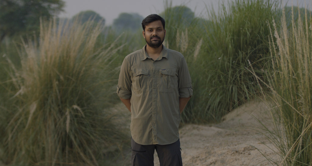 संरक्षणवादी गौरा चंद्र दास एक प्राकृतिक परिवेश में लंबी घासों से घिरे एक कच्चे रास्ते पर खड़े हैं।