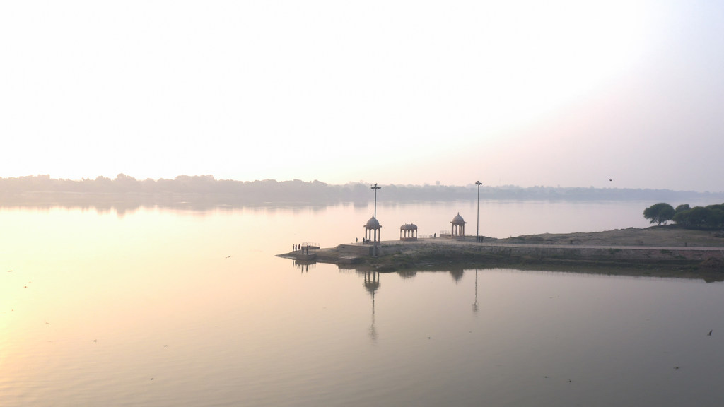 भारत में गंगा नदी पर सूर्योदय, नदी के किनारे मंडपों और दीपक स्तंभों से सुसज्जित एक घाट के साथ।