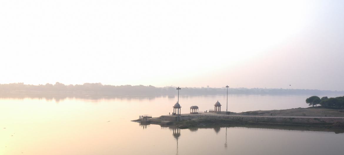 भारत में गंगा नदी पर सूर्योदय, नदी के किनारे मंडपों और दीपक स्तंभों से सुसज्जित एक घाट के साथ।