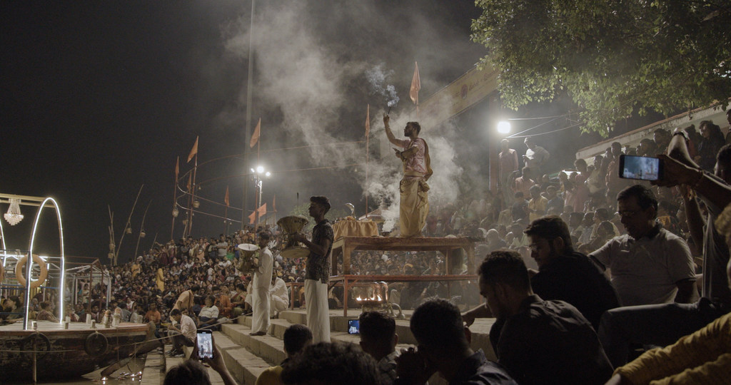 भारत के वाराणसी में गंगा नदी के किनारे एक चबूतरे पर पुजारी एक अनुष्ठान कर रहे हैं, और रात के समय नदी किनारे भारी भीड़ जमा है।
