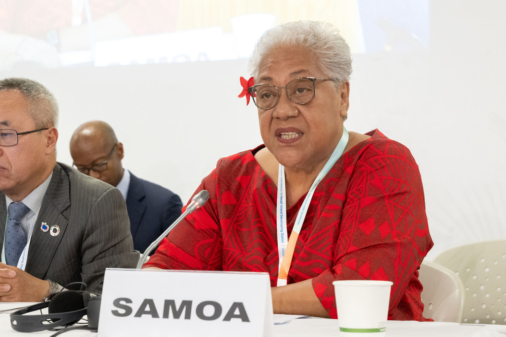 A woman with white hair and glasses, wearing a red patterned dress and a red flower in her hair, speaks at a conference table with a 'SAMOA' nameplate.