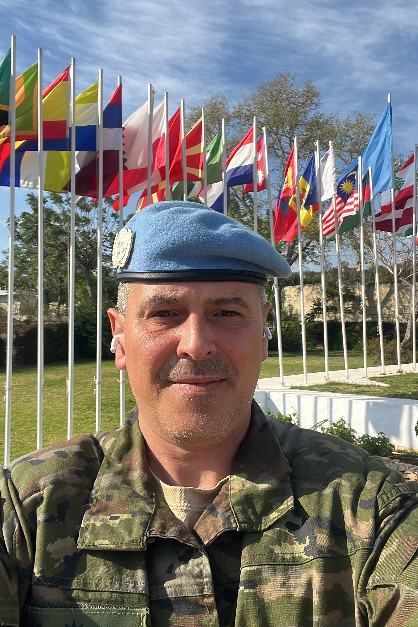 Un miembro español de las fuerzas de paz de la ONU , con boina azul y uniforme de camuflaje, posa frente a una hilera de banderas internacionales.