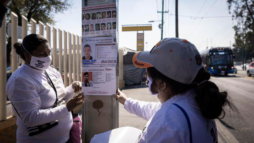 Dos mujeres están colocando carteles de personas desaparecidas en un poste de metal en una calle mexicana.
