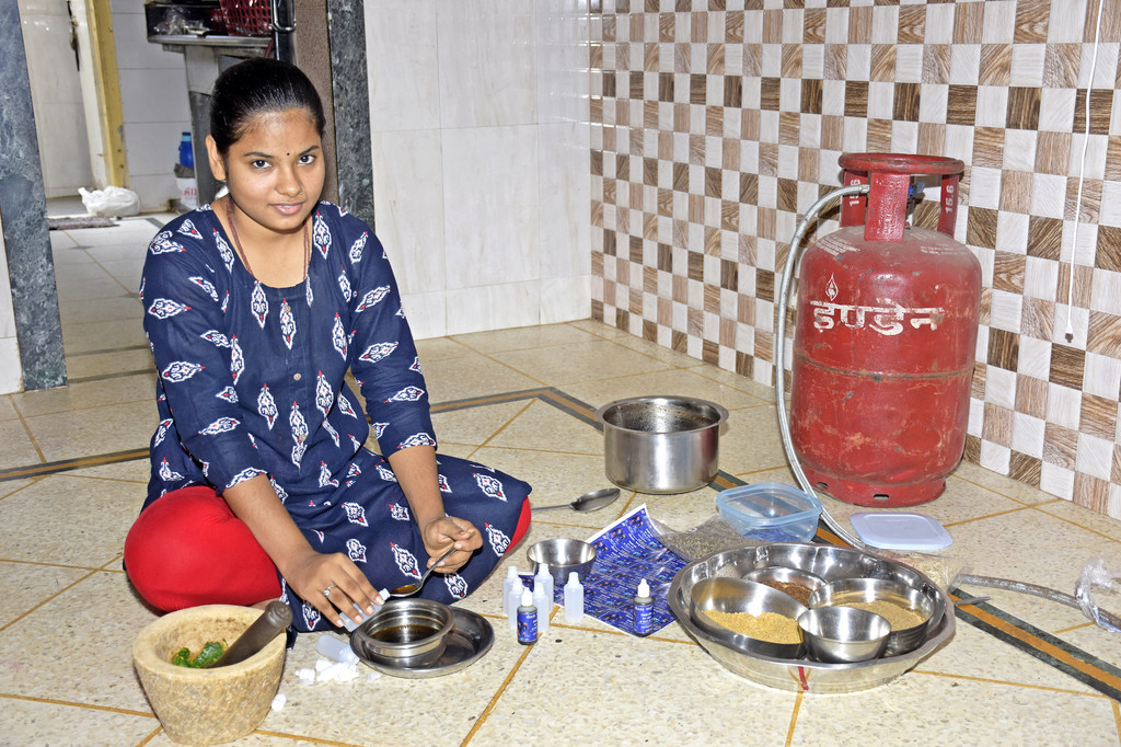 नीले रंग के पैटर्न वाले कुर्ते में एक भारतीय महिला फर्श पर बैठी है और ओखली और मूसल से आयुर्वेदिक औषधि तैयार कर रही है, उसके चारों ओर सामग्रियां और बर्तन रखे हुए हैं।