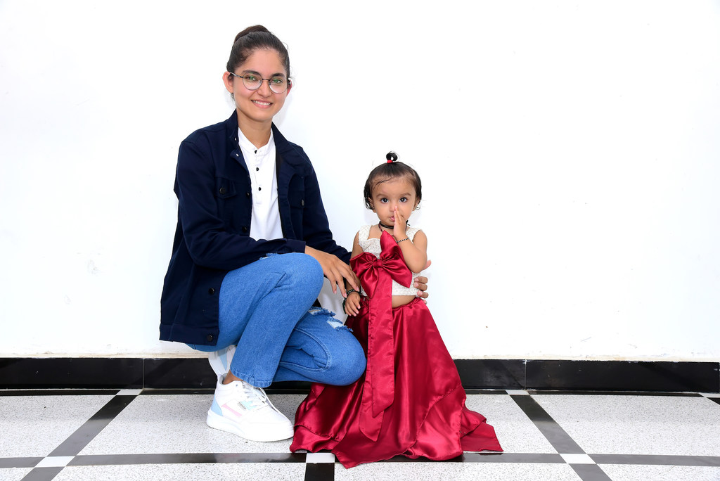 एक भारतीय महिला और बच्चा तस्वीर खिंचवा रहे हैं। महिला ने नेवी ब्लू जैकेट और नीली जींस पहनी है, जबकि बच्चे ने लाल रंग की ड्रेस पहनी है जिस पर एक बड़ा सा रिबन लगा है।
