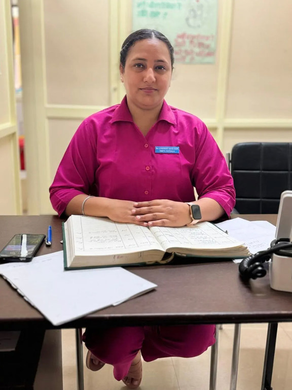 भारत के लुधियाना में एक कार्यालय में दाई लवप्रीत सैनी एक डेस्क पर बैठी हैं, उन्होंने गुलाबी रंग की वर्दी पहनी हुई है, जिस पर नाम का टैग और घड़ी लगी हुई है।