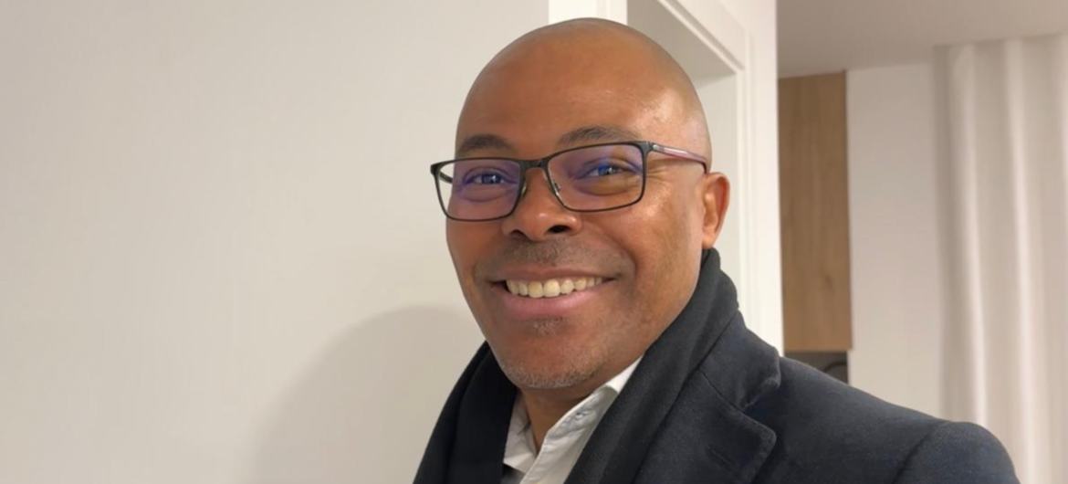 Retrato de um homem negro sorridente, de óculos, usando um cachecol e jaqueta escuros, em ambiente interno.