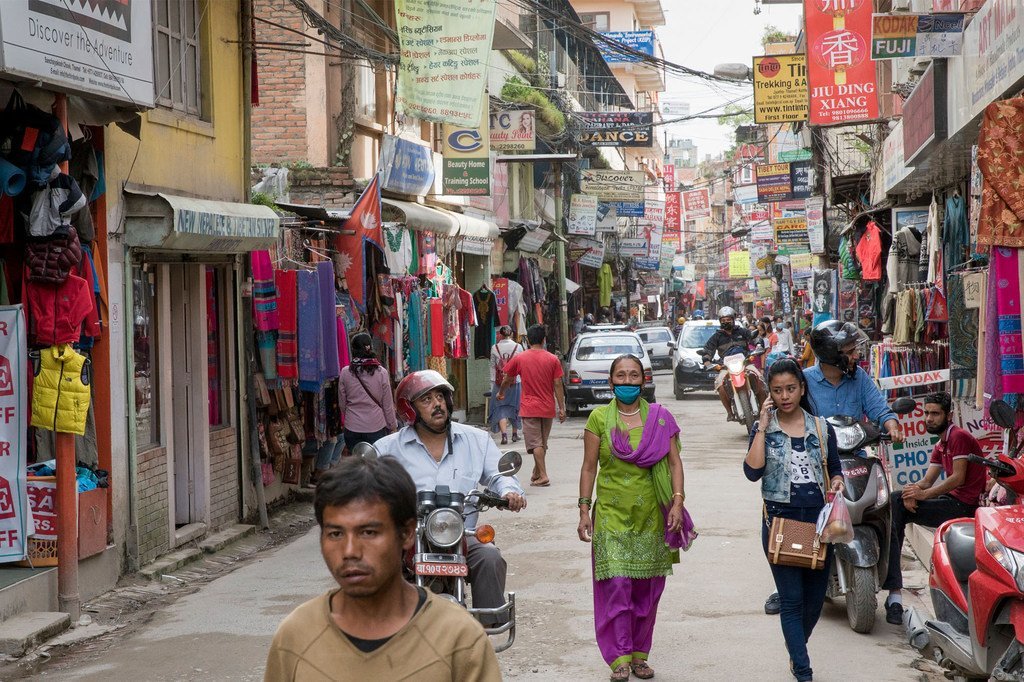 नेपाल के काठमांडू शहर की एक व्यस्त सड़क पर पैदल यात्री और मोटरसाइकिल सवार नजर आ रहे हैं, जिसके किनारों पर दुकानें और रंग-बिरंगे वस्त्र सजे हुए हैं।