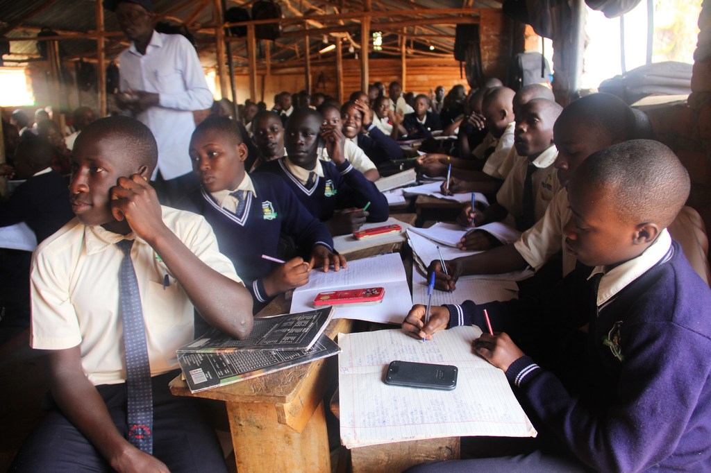 Wanafunzi waliovaa sare za shule wamekaa kwenye madawati ya mbao darasani, wakiandika kwenye madaftari kwa kalamu.
