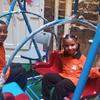 Watoto wawili wadogo waliovaa sare za shule za rangi ya chungwa wamekaa kwenye seti ya swing katika eneo la kuchezea la ndani.