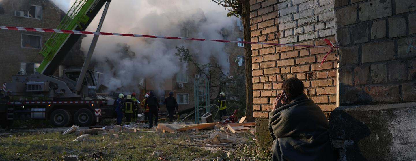 Des équipes de secours et une grue travaillent sur le site d'une frappe de missile sur un immeuble résidentiel à Kyiv, en Ukraine, tandis qu'un habitant est assis à proximité, enveloppé dans une couverture.