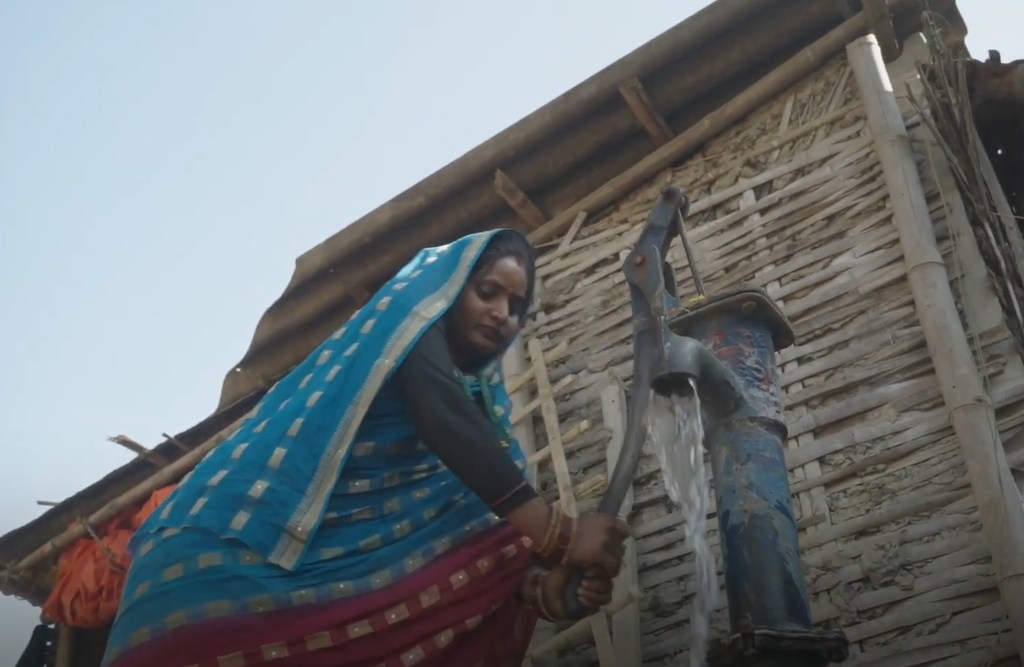 Mwanamke aliyevaa sari ya bluu na nyekundu akiendesha pampu ya maji nje ya nyumba ya vijijini huko Nepal.