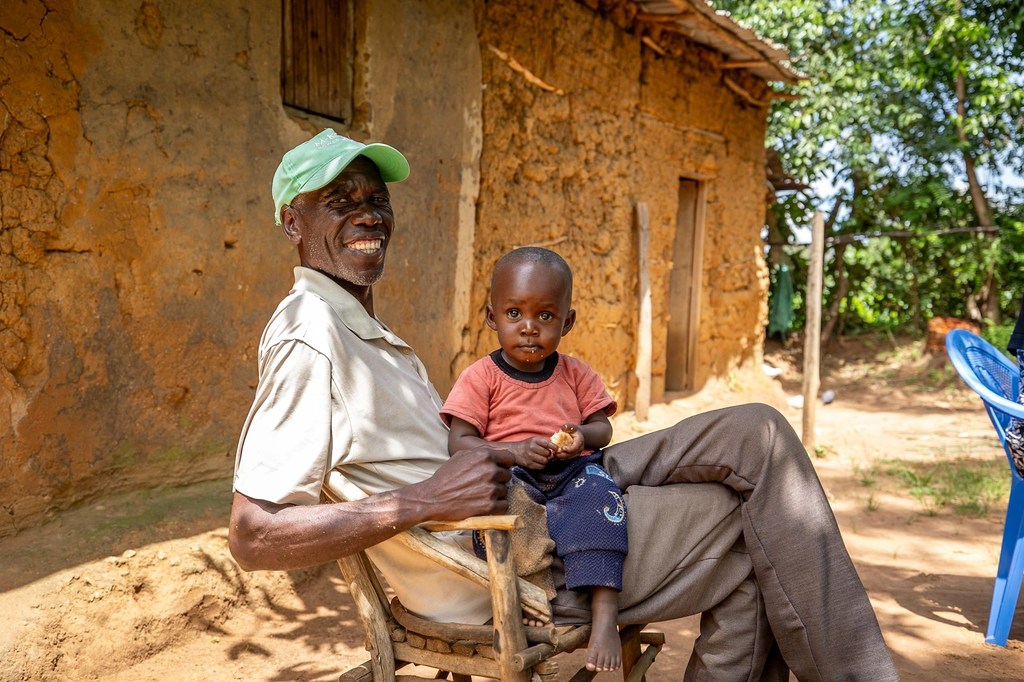 Mkulima Mkenya anayetabasamu anayeitwa Lusakia amembeba mtoto mdogo huku akiwa ameketi kwenye kiti cha mbao nje ya nyumba ya udongo huko Bungoma.