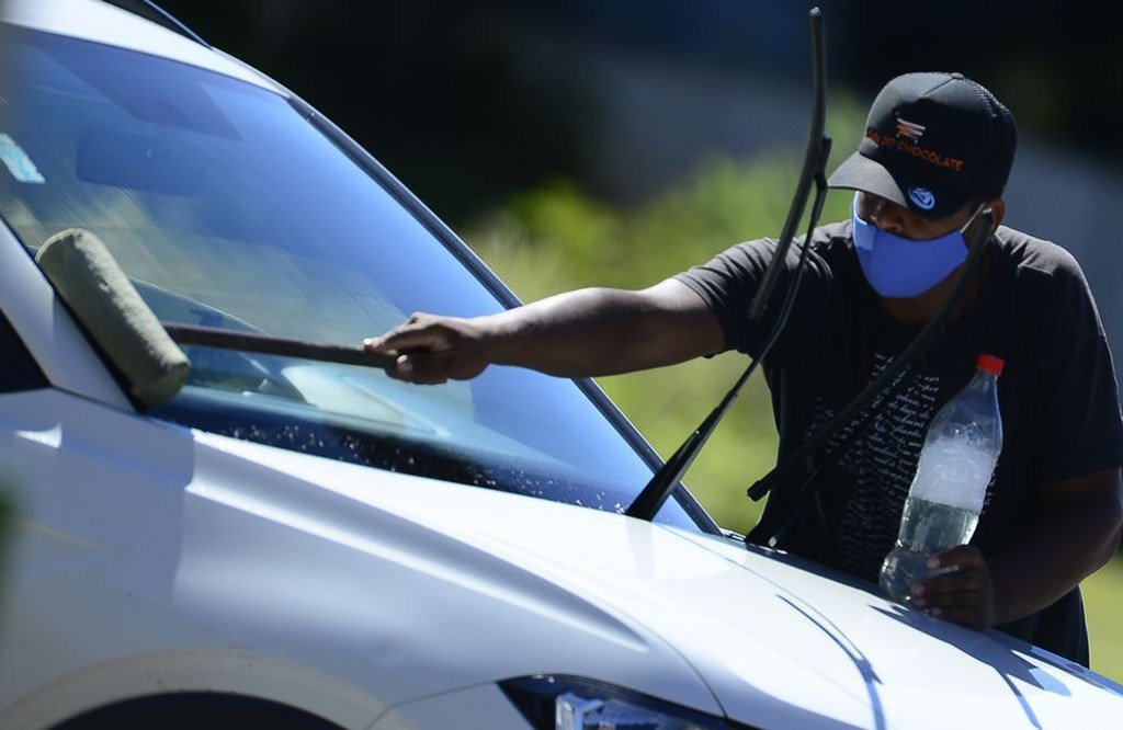 En Brasil, un hombre, con mascarilla y gorra, limpia el parabrisas de un coche blanco.