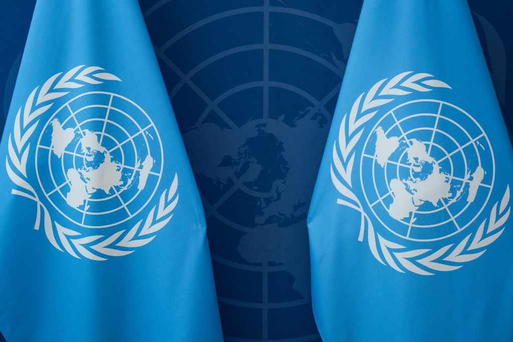 Two United Nations flags with the UN emblem displayed in front of a wall featuring a large UN emblem.