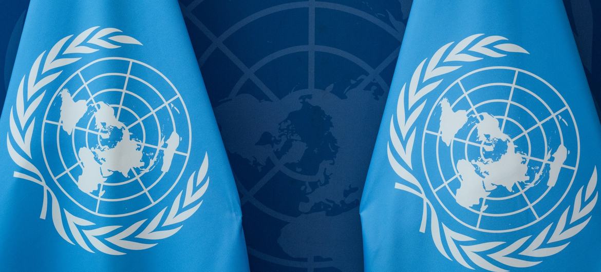 Two United Nations flags in front of a UN emblem on the wall behind.