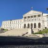 Um grande palácio neoclássico com uma imponente escadaria e colunas, com a bandeira portuguesa hasteada no topo.