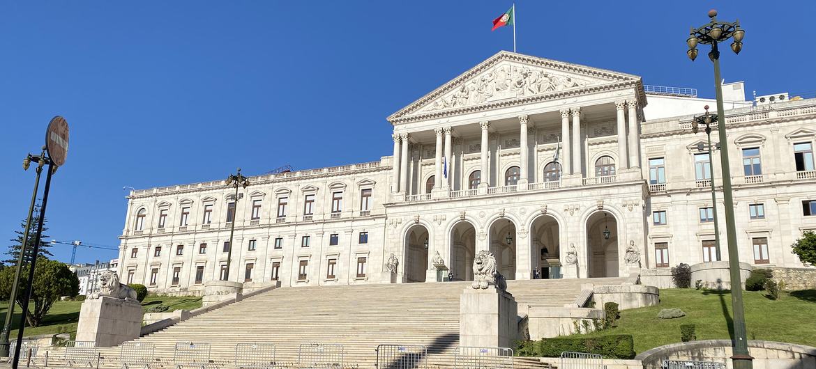 Um grande palácio neoclássico com uma imponente escadaria e colunas, com a bandeira portuguesa hasteada no topo.