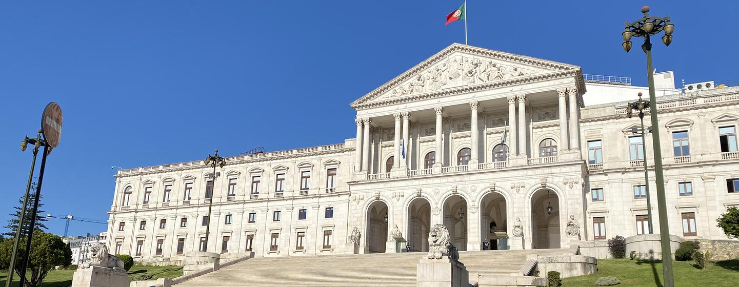 Um grande palácio neoclássico com uma imponente escadaria e colunas, com a bandeira portuguesa hasteada no topo.