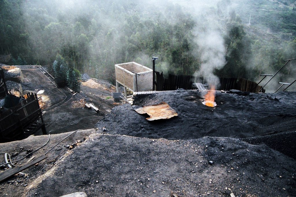 Vista aérea de uma mina de carvão em Samaca, Colômbia, mostrando uma grande pilha de carvão com uma chama de metano acesa no topo, cercada por uma paisagem florestal.
