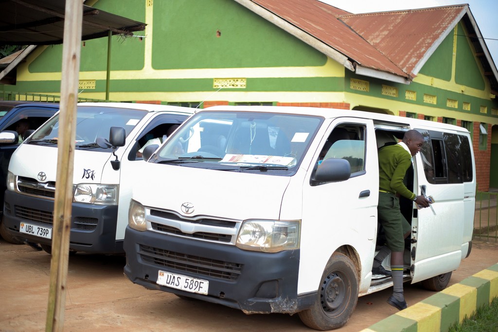 Mwanamume aliyevaa sare ya kijani anaingia kwenye basi dogo jeupe la Toyota lililokuwa limeegeshwa mbele ya jengo la shule ya kijani huko Kampala, Uganda. Gari hilo ni sehemu ya usafiri unaotolewa na UNICEF kwa wanafunzi wa Shule ya Msingi ya Green Valley.