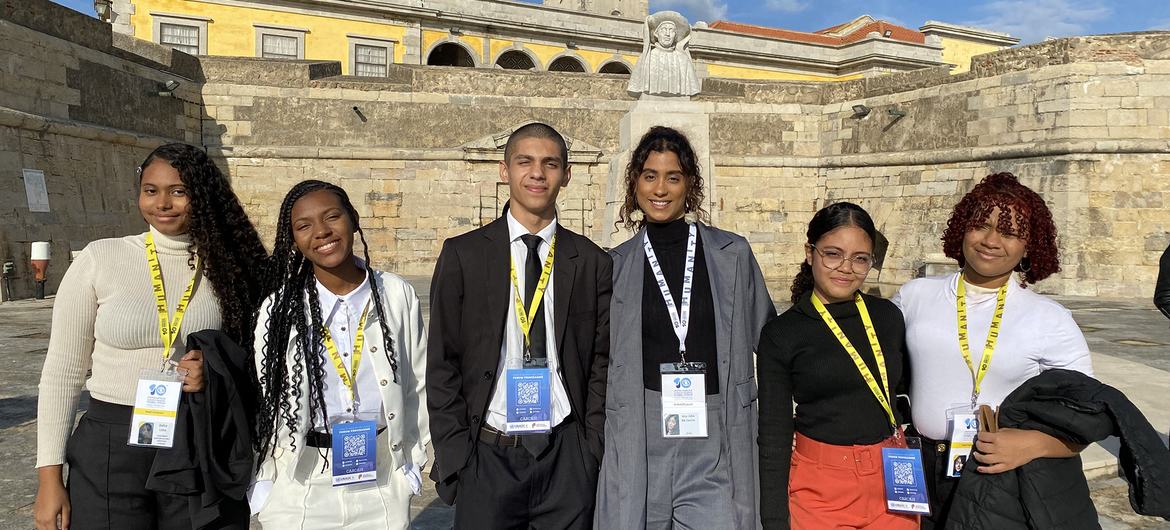 Um grupo de seis estudantes brasileiros, usando crachás e cordões do evento, posa para uma foto em frente a um prédio histórico de pedra.