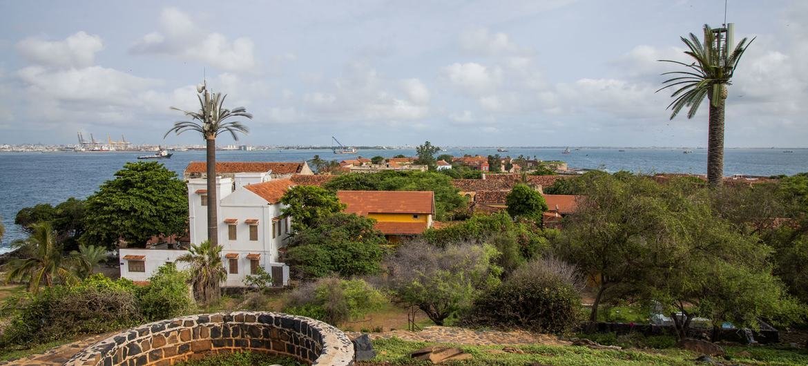 Vue de l'île de Gorée avec le port de Dakar en arrière-plan, mettant en valeur des bâtiments de style colonial, des palmiers et un puits de pierre au premier plan.