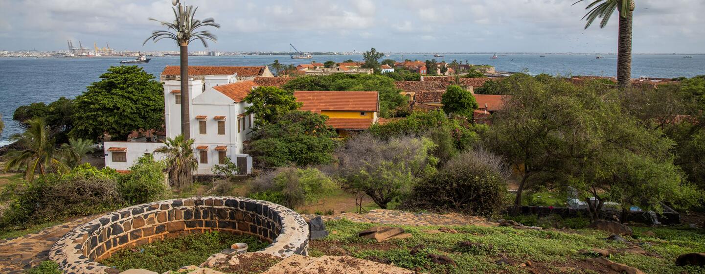 Vue de l'île de Gorée avec le port de Dakar en arrière-plan, mettant en valeur des bâtiments de style colonial, des palmiers et un puits de pierre au premier plan.