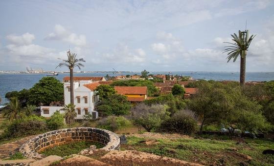 Vue de l'île de Gorée avec le port de Dakar en arrière-plan, mettant en valeur des bâtiments de style colonial, des palmiers et un puits de pierre au premier plan.