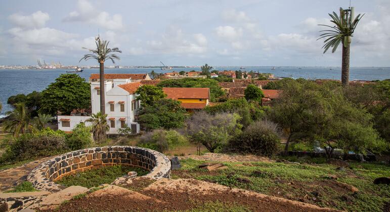 Vue de l'île de Gorée avec le port de Dakar en arrière-plan, mettant en valeur des bâtiments de style colonial, des palmiers et un puits de pierre au premier plan.