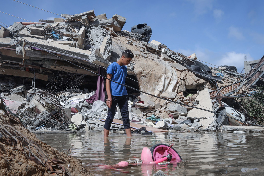 Un niño palestino con una camisa azul camina por una calle inundada junto a un montón de escombros de un edificio destruido en la ciudad de Gaza.
