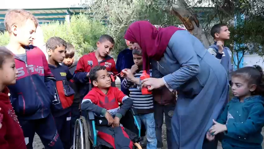 A woman in a hijab distributes water to a group of children, including one in a wheelchair, at an outdoor event in Gaza.