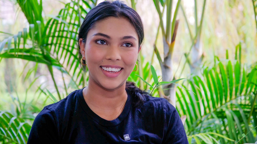 Un retrato de la activista climática trinitense Priyanka Lalla sonriendo, con un fondo de hojas de palmera verdes.