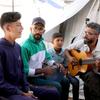 أحمد أبو عمشة يعزف على الغيتار لمجموعة من الصبية في خيمة، بينما يعزف رجل آخر على آلة إيقاعية.