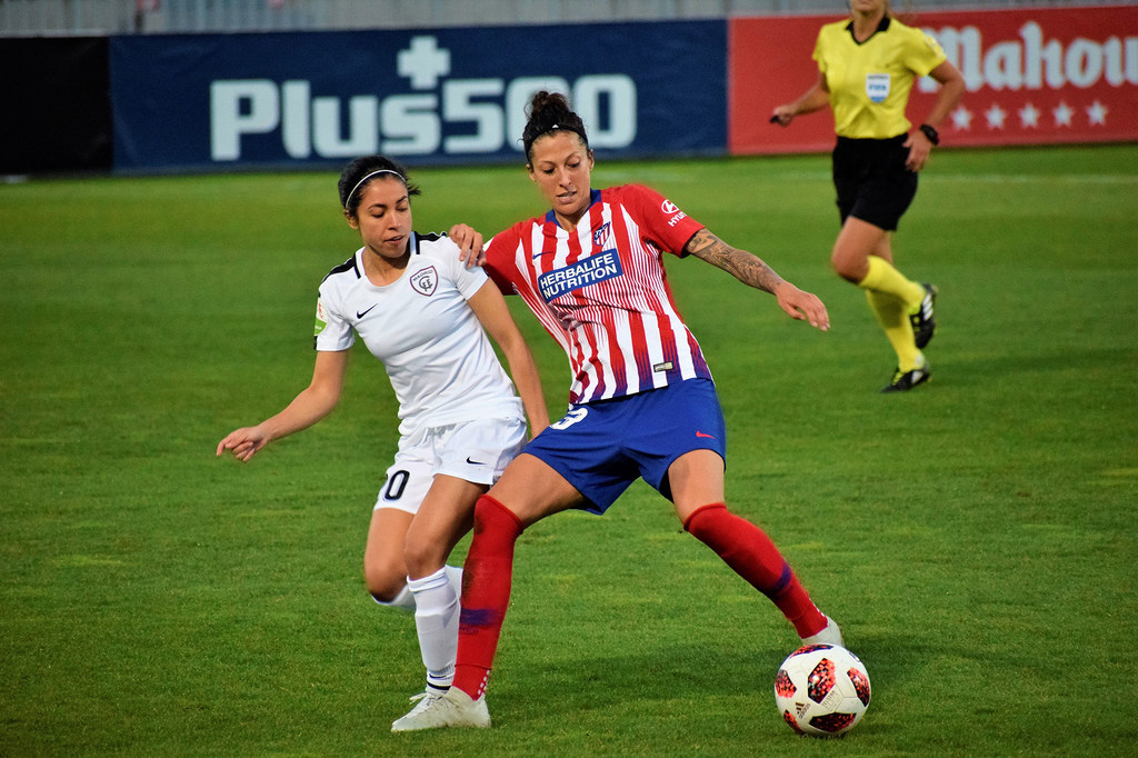 Dos jugadoras de fútbol femenino compiten por el balón durante un partido, con una de ellas controlándolo cerca de la línea de banda.