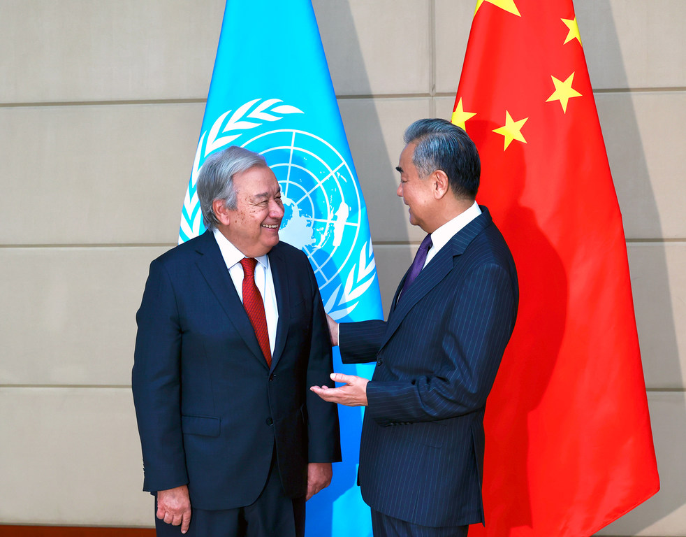 El Secretario General de la ONU, António Guterres, y el Ministro de Relaciones Exteriores de China, Wang Yi, se ven conversando, de pie frente a las banderas de la ONU y de China.