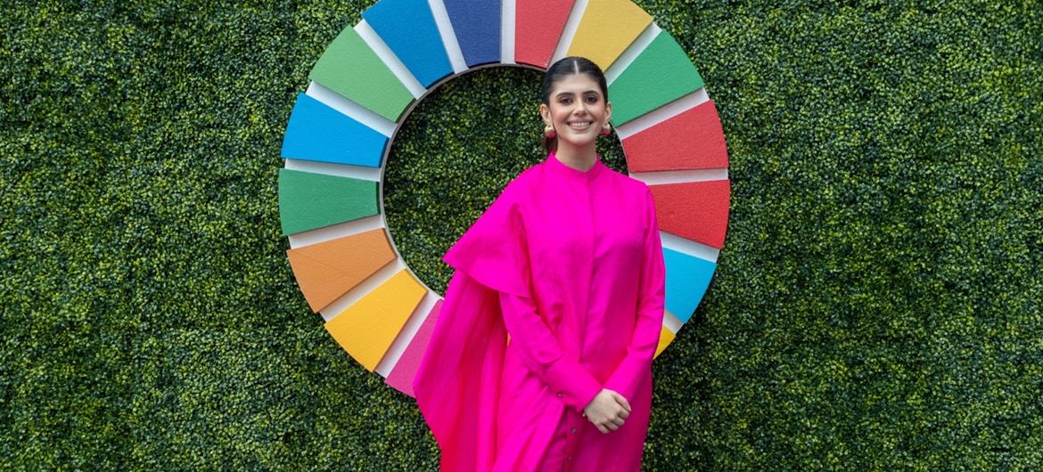 गुलाबी रंग की पोशाक पहने भारतीय महिला संजना सांघी एक हरी दीवार के सामने मुस्कुराते हुए खड़ी हैं, जिस पर एसडीजी का बड़ा लोगो और 'एसडीजी मी... जोन' लिखा हुआ है।